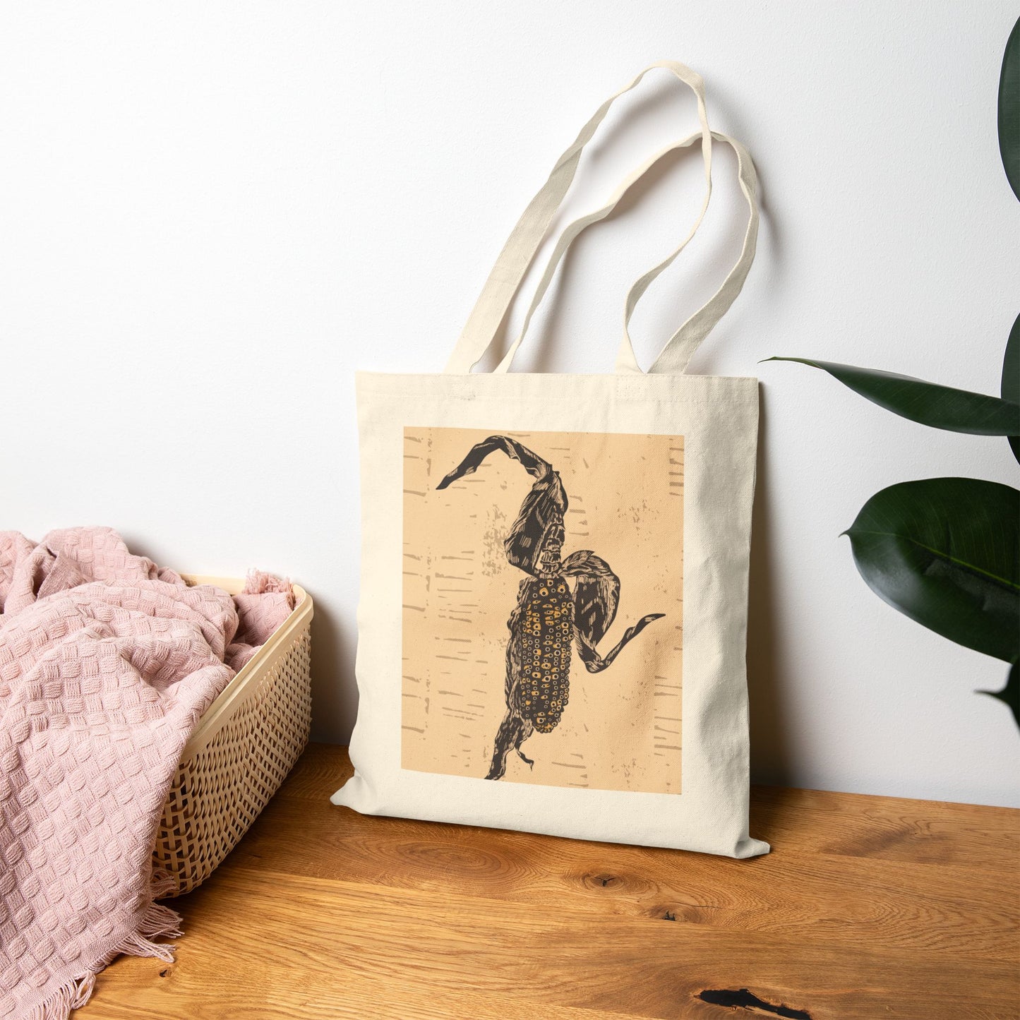 Tote bag with a scorpion design on a wooden surface next to a pink blanket and plant.